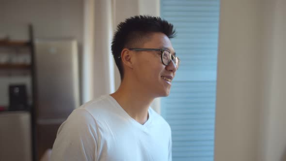 Young Asian Man in Glasses and Pajama Standing at Window at Home Looking Away