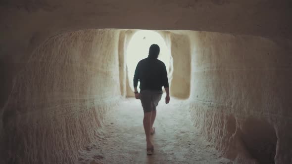 Unrecognizable man walking out of a cave, light at the end of the tunnel concept, back view dolly ou