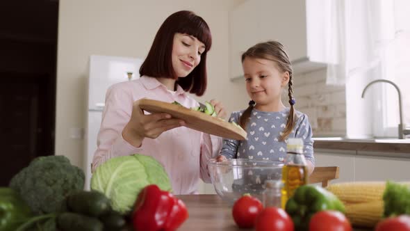 Cute Daughter Prepare Lunch with Mom Throwing Sliced Cucumbers in Glass Bowl in Modern Kitchen Room