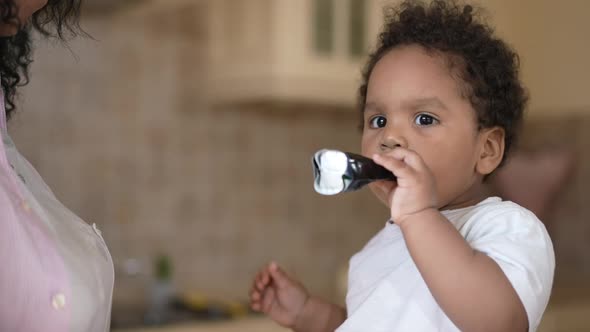 Zoom in to Serious Toddler Boy Eating Tubed Children Food Sitting in Kitchen with Unrecognizable