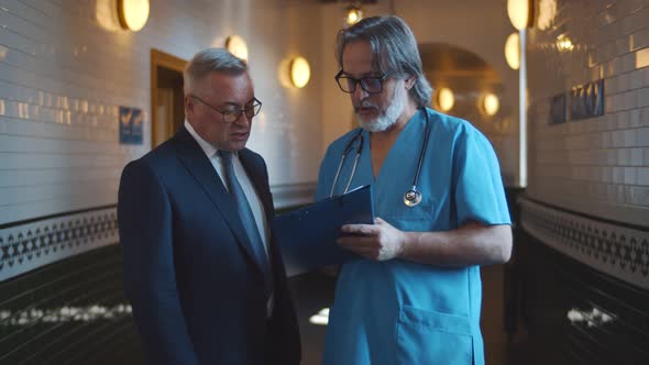 Senior Male Doctor in Uniform Discussing Diagnosis with Man Patient Looking at Clipboard