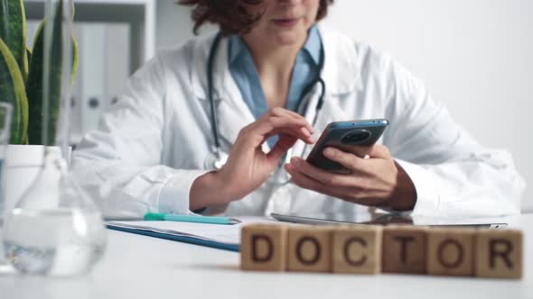 Girl Doctor Holding A Phone. Female Medic Flipping Through The Phone While Sitting At The Workplace