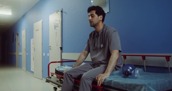 A Doctor Sits on a Medical Gurney and Rubs His Head in Empty Hospital Corridor