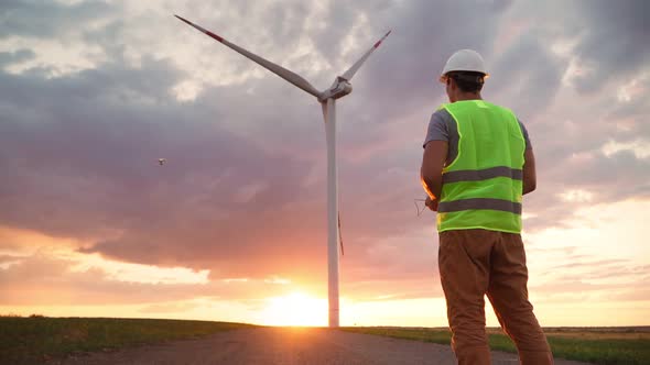 Man Engineer in uniform and helmet holding remote controller flying drone working at wind turbine