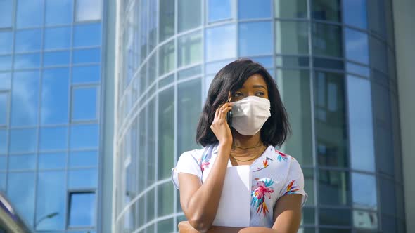 African American Young Business Woman in Protective Mask Is Talking on Phone Outside Near Modern