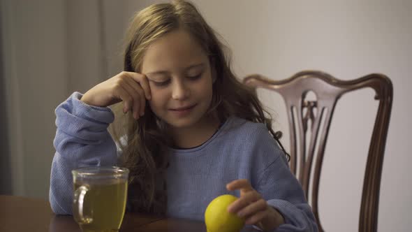 Close-up of Little Girl Holding Lemon While Drinking Herbal Tea. Child Sitting at the Table at Home