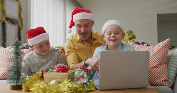 Caucasian father and two sons wearing santa hats having a videocall on laptop during christmas