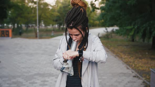 Portrait of Young Pretty Woman with Dreadlocks Smoking Marijuana with a Bong in Park