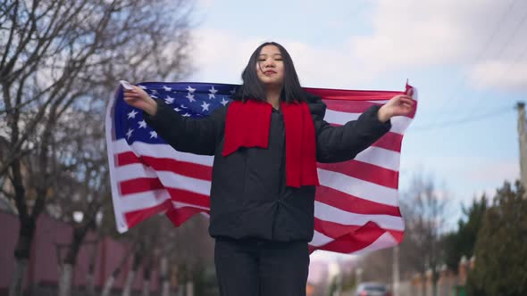 Portrait of Confident Patriotic Asian Woman Wrapping in American Flag Looking at Camera Smiling