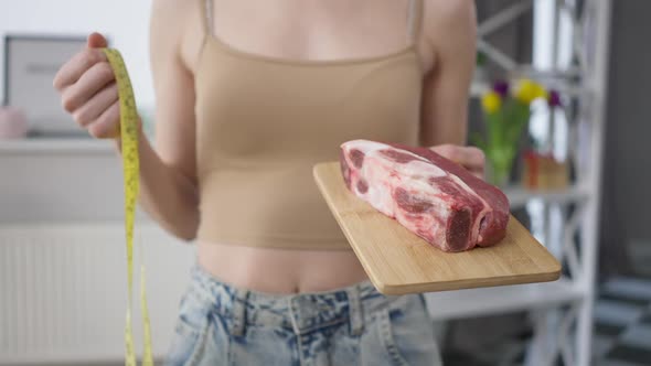Unrecognizable Fit Slim Woman Holding Raw Beef Steak and Measuring Tape Standing Indoors