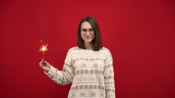 A Young Woman in a White Sweater Holds a Sparkler in Her Hand. Shooting in the Studio on a Red