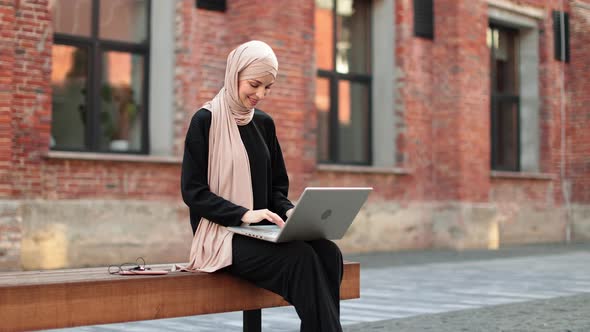 Pretty Smiling Muslim Business Woman in Hijab Sitting on Bench and Using Laptop During Video Chat