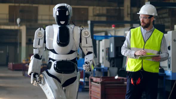 Factory Worker Checks a Walking Cyborg, Using a Tablet.