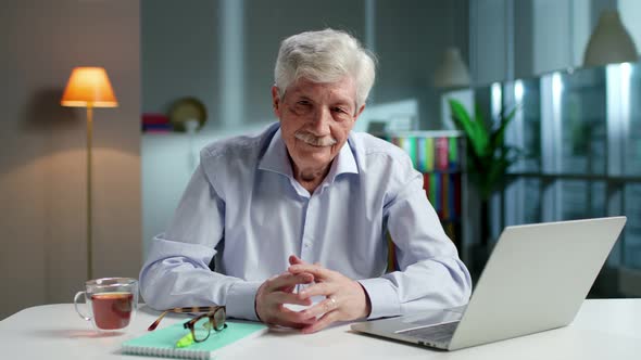 Portrait of Senior Man Take Off Glasses and Looking at Camera Sitting at Desk