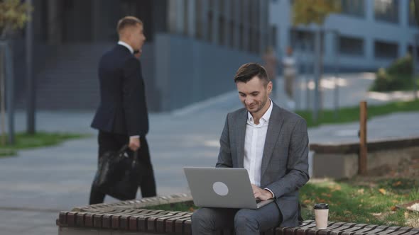 Young Businessman in the City Sits on Bench Takes a Laptop and Works