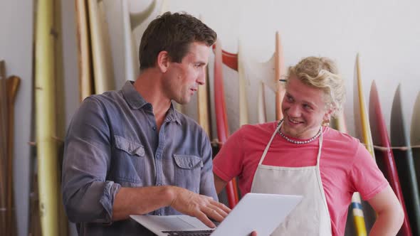 Two Caucasian male surfboard makers working on projects using a laptop computer