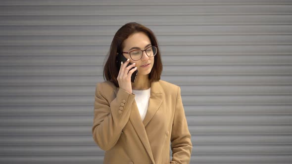 A Young Woman Stands on a Gray Background and Speaks on the Phone. Girl in a Coat.