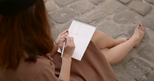 Girl Artist Sits on the Sidewalk and Paints in a Notebook