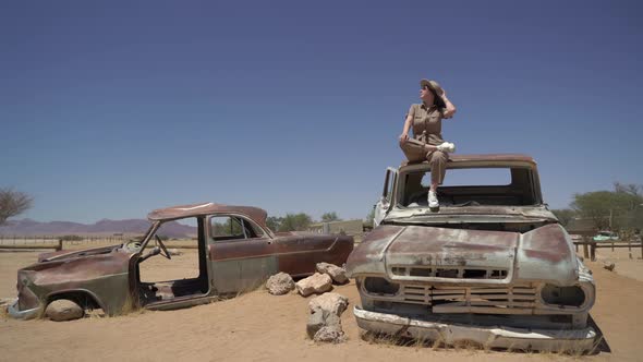 Young Woman in Safari Overalls Sits on the Rooftop of Abandoned Old Rusty Car