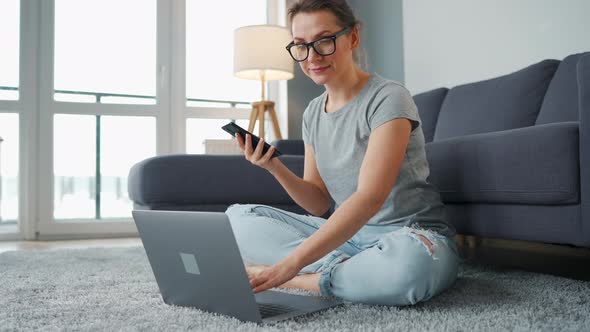 Casually Dressed Woman Sitting on Carpet with Laptop and Smartphone and Working in Cozy Room