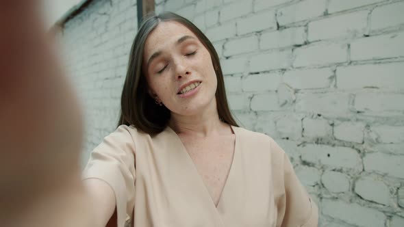 Portrait of Cute Young Lady with Freckles Chatting and Waving Hand During Video Call Outdoors