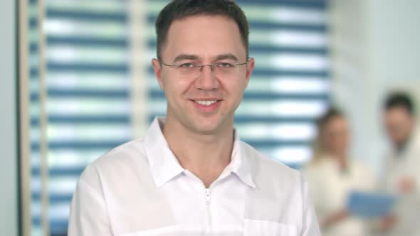 Male Doctor in Glasses Smiling at Camera While Medical Staff Working on the Background