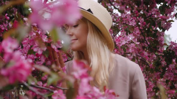 Portrait Romantic of a Blonde Woman in Purple Dress and Hat with Long Hair Standing By a Flowering