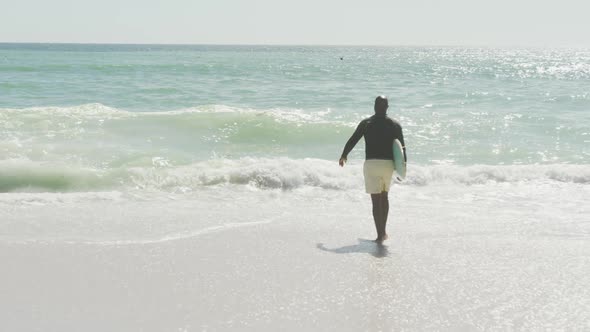 Senior african american man walking with surfboard on sunny beach