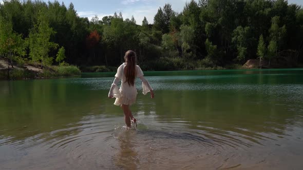 One Young Woman in a White Dress Goes and Wets Her Feet on a Wild Forest Lake in Summer.