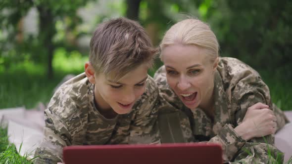 Carefree Military Woman and Teenage Boy Talking Laughing Surfing Internet on Laptop Lying on Sunny