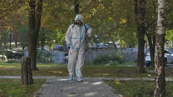 Virologist in a White Protective Suit Disinfects Paving Slabs in a Public Park. The Man Uses a High