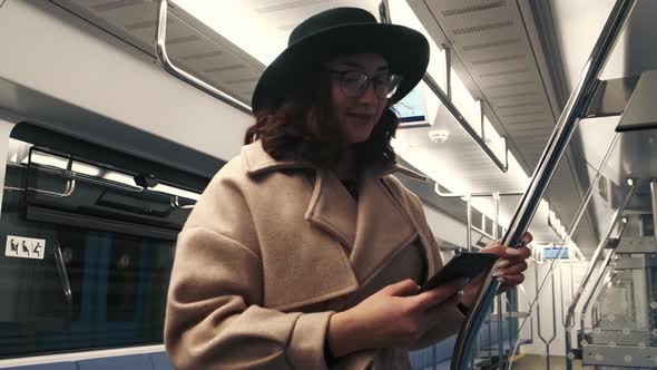 A Pretty Young Girl in a Coat with a Black Hat is Sitting on the Phone While in a Subway Car