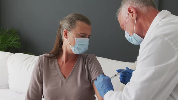 Caucasian senior male doctor wearing face mask giving covid-19 vaccine to woman at home