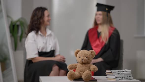 Teddy Bear and Scientific Books on Table with Blurred Mature Mother and Graduate Daughter Talking at