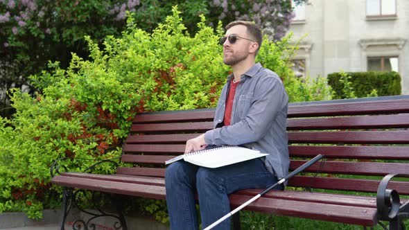 Young Blind Man Sitting on Bench in City Park and Reading a Braille Book