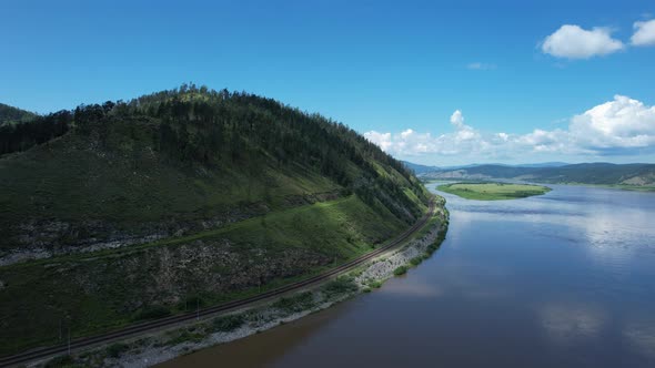 Flying Above a Glacial River in Siberia Russia with Green Mountains and Railway Track in the