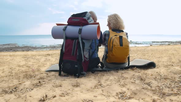 Back View of Senior Backpack Couple Sitting on Beach Enjoying Seascape