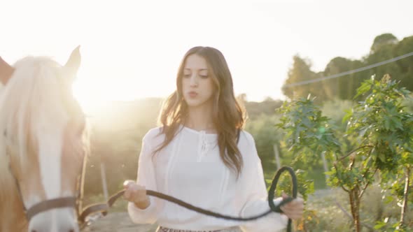 Girl in White Dress Walks with Horse on a Leash in the Ranch