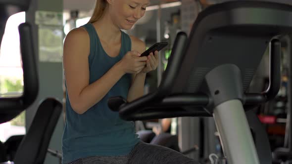 A Young Woman at the Gym on an Exercise Bike Holding the Phone in Front of Her Eyes in Her Hands