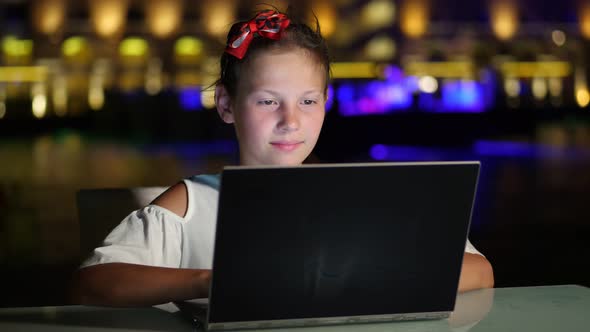 Cute Teen Girl Uses Laptop, Sitting at Table on Open Terrace or Balcony, Against Background of Night