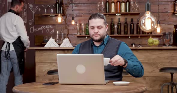 Handsome Man with Long Hair Looking at His Laptop Screen and Drinks Coffee