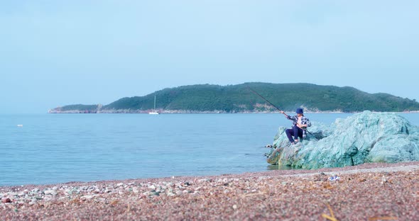 Fisherman Sits on Rocks and Throws Fishing Rod Into Water