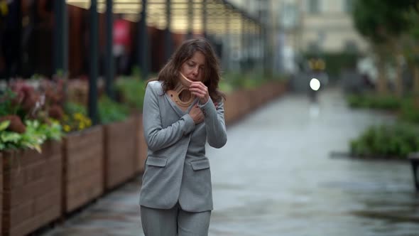 Sad Woman with Orthopedic Cervical Collar on Neck Is Walking on Street in City at Rainy Day