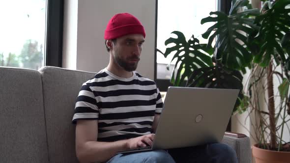 Modern, young man working on a laptop, sitting on a sofa in a cafe or at home.