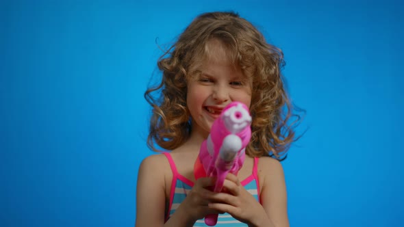 Little Blond Curly Girl in Swimsuit with Pink Water Gun Smiles in the Studio