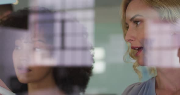 Diverse businesswomen in discussion at work, caucasian woman writing notes on glass wall