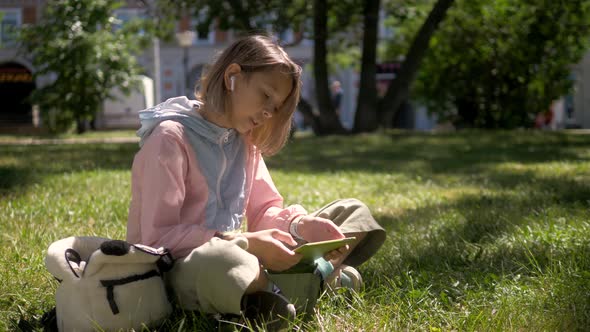 Little Girl Child Kid Sitting on Grass in Park Looking Smart Watch and Put Airpods. Smart Watch for