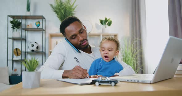 Black-Skinned Dad Talking on Phone, Making Notes Into Notebook and Watching at His Small Son