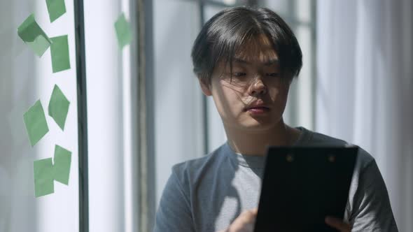 Portrait of Focused Asian Young Man Analyzing Paperwork in Sunlight Indoors