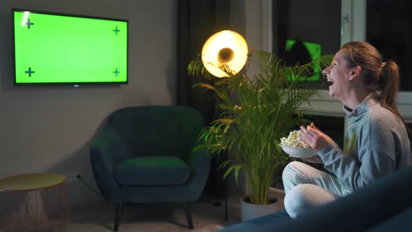 Rear View of a Woman Sitting on a Sofa in the Living Room in the Evening and Watching a Green TV